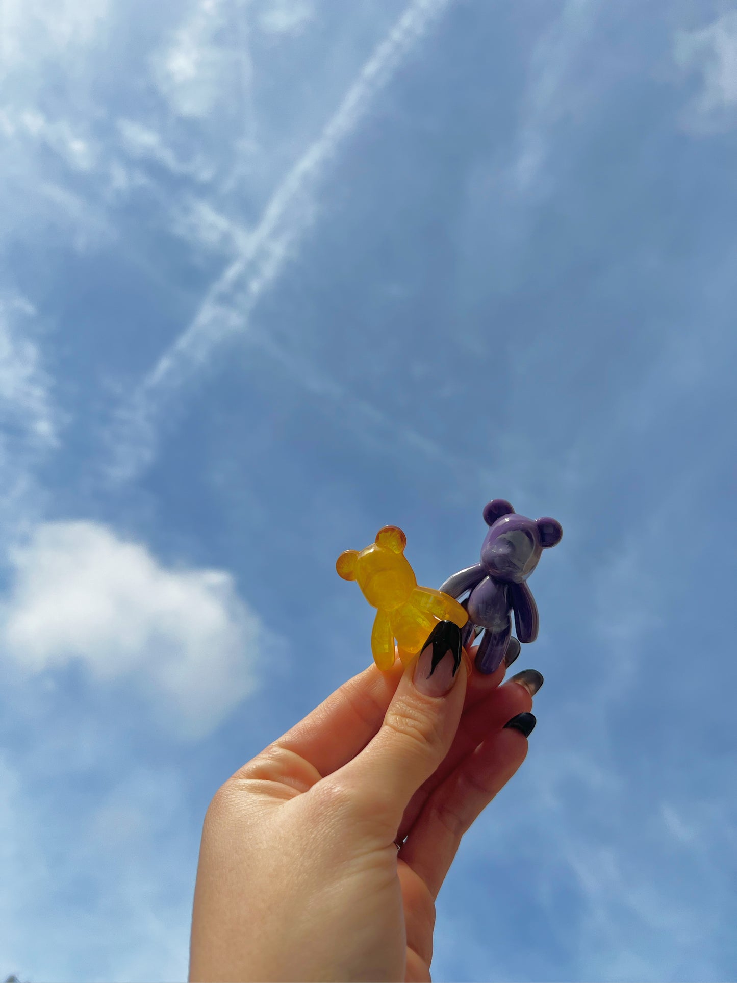 Yellow see through bear and purple marble bear against a bright blue sky in an adult hand with black painted nails.